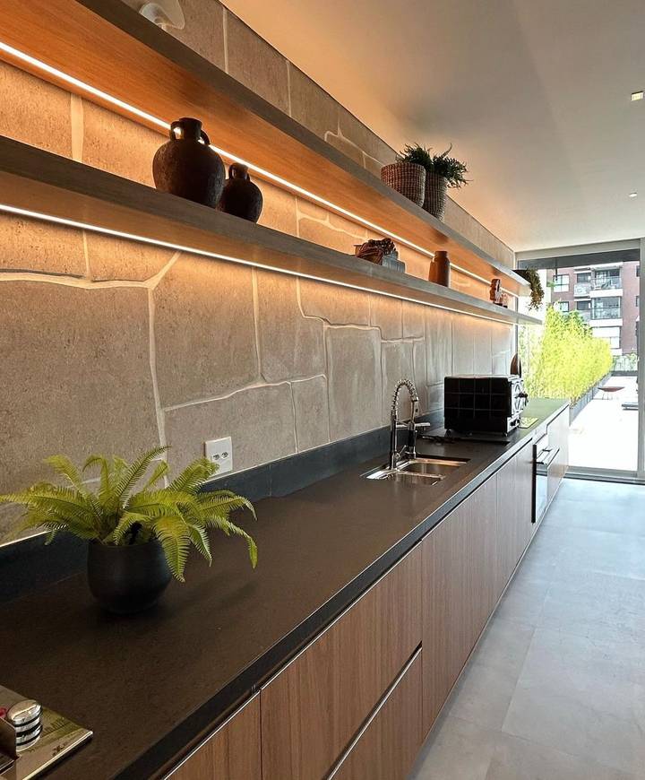 Cozinha gourmet com bancada em pedra escura, armários de madeira e prateleiras iluminadas